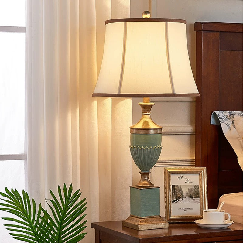 Sage green and antique-gold table lamp with a cream fabric shade on a bedside table.