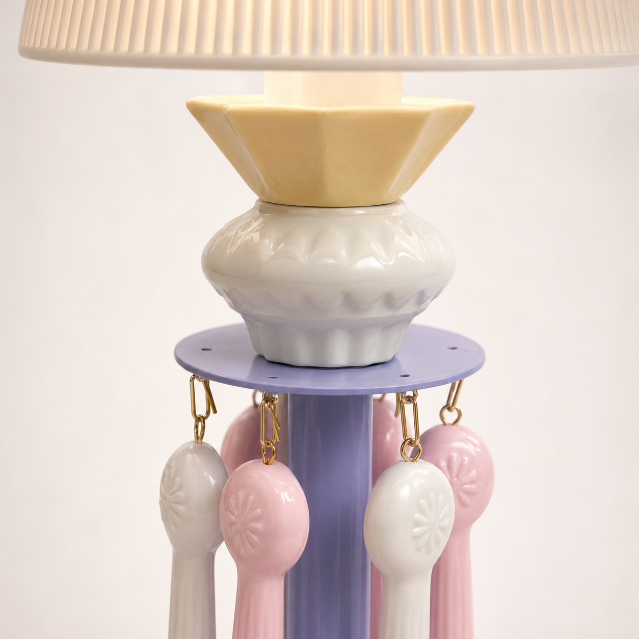 A decorative table lamp with a pleated off-white shade, a pale yellow geometric collar, a white textured ceramic pot, a lavender circular platform, and hanging pastel pink and off-white spoon-shaped accents.