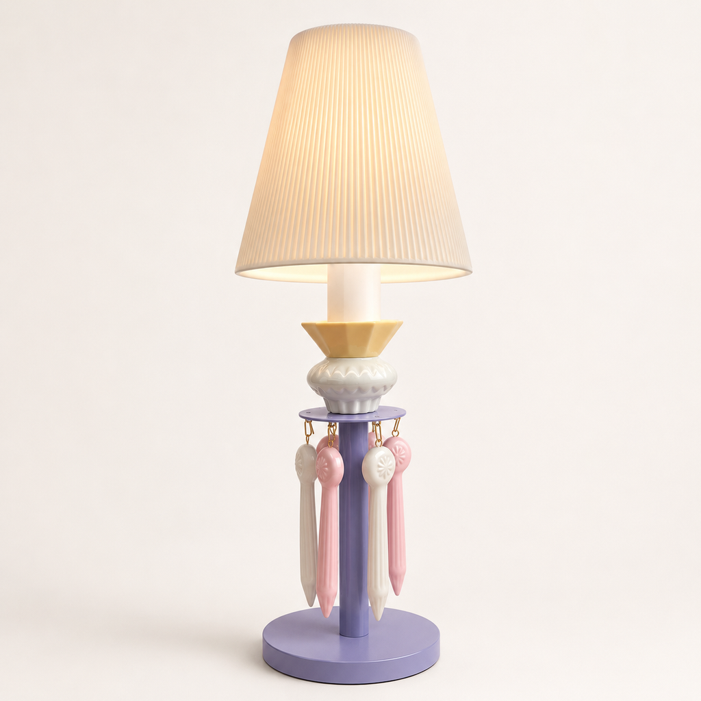A decorative table lamp with a pleated beige fabric shade, a lavender-purple base, a pale yellow collar, and hanging pink and white ribbons, shown against a plain light background.