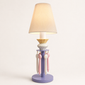 A decorative table lamp with a pleated beige fabric shade, a lavender-purple base, a pale yellow collar, and hanging pink and white ribbons, shown against a plain light background.