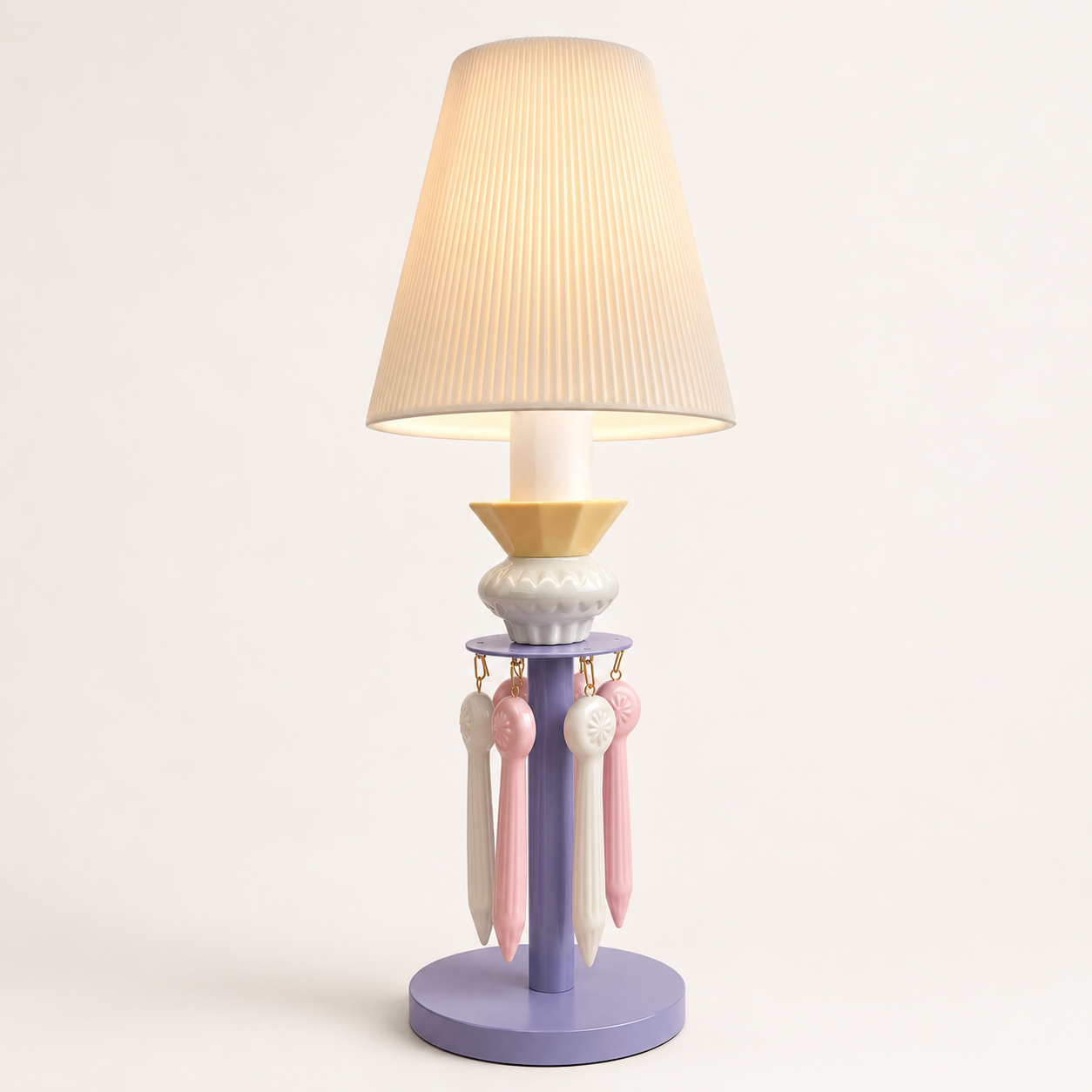 A decorative table lamp with a pleated beige fabric shade, a lavender-purple base, a pale yellow collar, and hanging pink and white ribbons, shown against a plain light background.