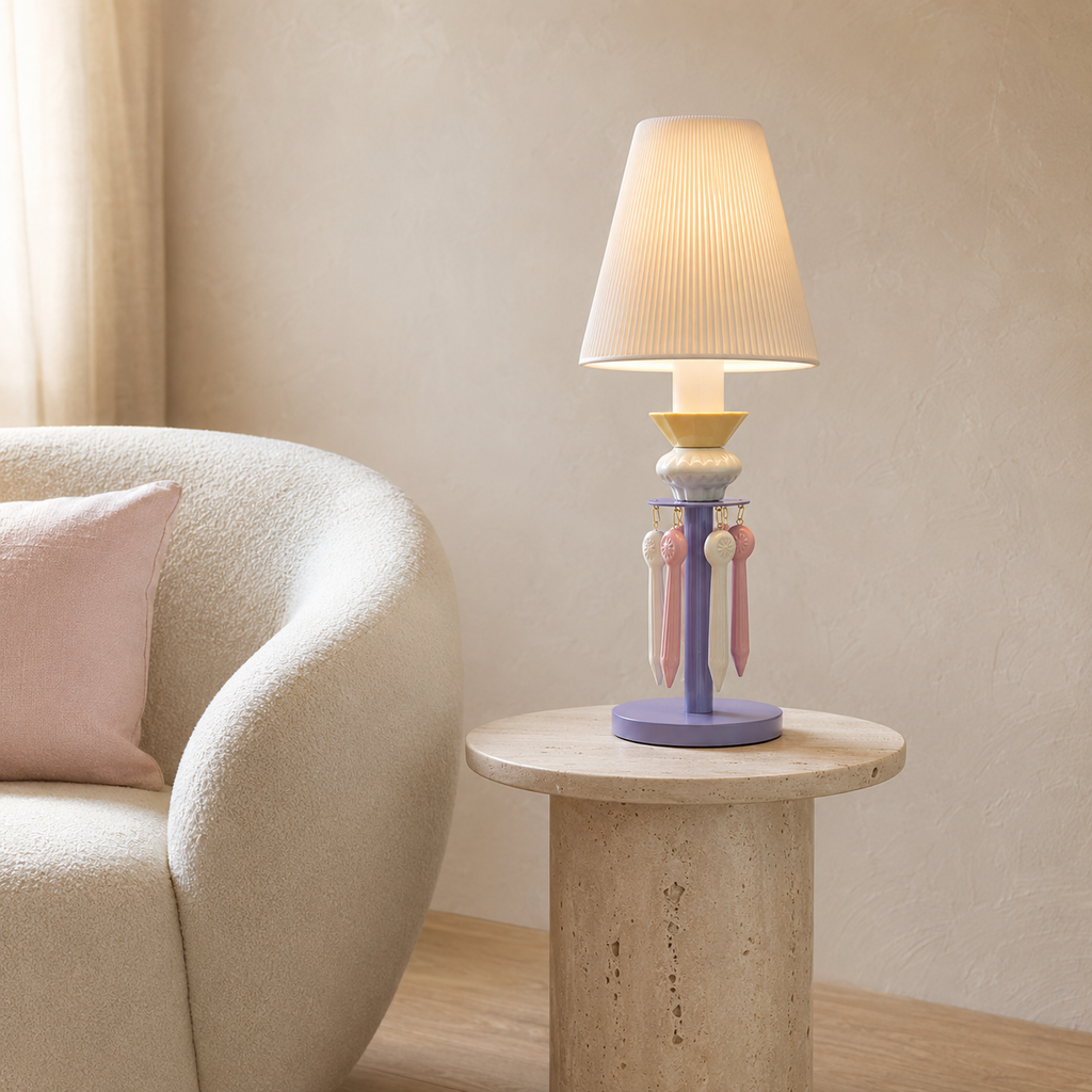 A decorative table lamp with a pleated white shade, a lavender base, and pastel pink and yellow hanging accents, placed on a round stone side table next to a cream upholstered armchair in a softly lit room.