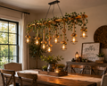 A rustic wooden dining room scene with a large hanging chandelier made from a wooden frame, green ivy, rope details, and multiple glowing wire cage pendant lights. A dining table, chairs, window, and wall decor are visible in the room.